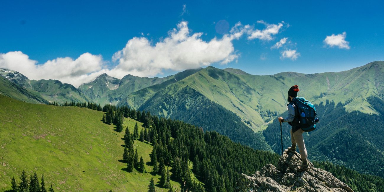 Adventurous hiker enjoying breathtaking views of lush green mountains in Romania under a clear blue sky.